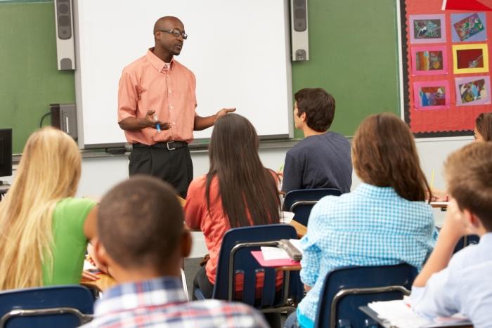 A classroom full of students sitting in their seats facing the front of the classroom to a teacher talking.