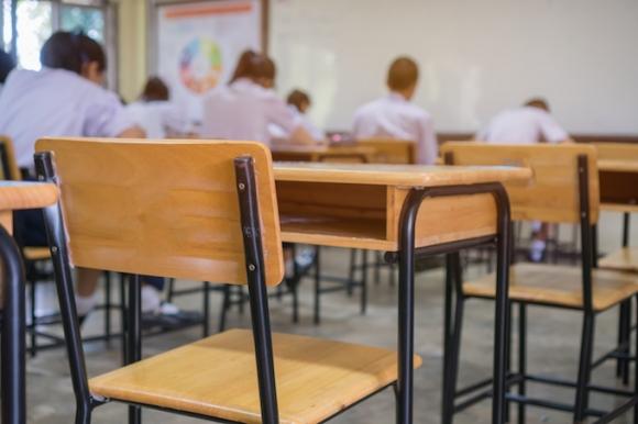 A focused image of an empty chair desk, while in the background are students writing while in their desks.